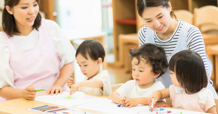 保育施設での光景。3人のおこさんがテーブルに座りなにか作業をしているところを二人の保育士がほほえみながら見守っている。