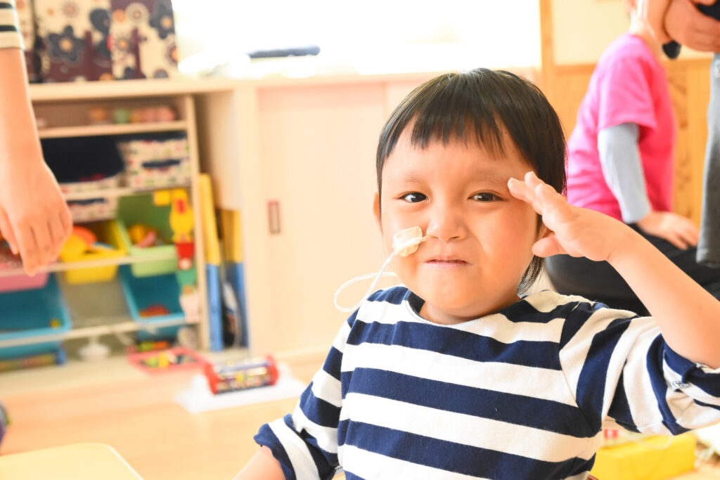 経鼻チューブをつけたこどもが、障害児保育園ヘレンの保育室で笑顔で手をあげている。医療的ケア児の日常の一場面。